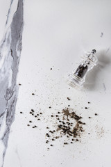 Black peppers peppercorns and ground powder from transparent pepper mill over white marble texture...