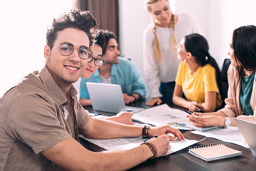 two multicultural businessmen in eyeglasses looking at camera while their partners having discussion behind at modern office