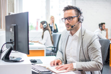 Serious man wearing formal clothes and headset looking at computer screen in bright office