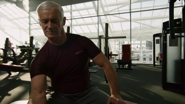 PAN of elderly man with grey hair sitting on gym bench and training arms with dumbbell biceps curl exercise