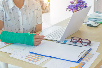 injured woman with green cast on hand and arm working on laptop in office, focus on broken hand