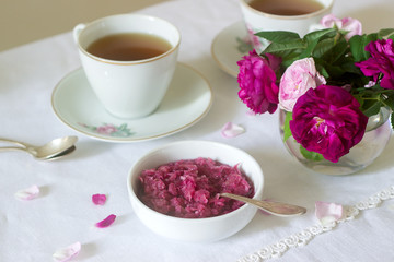 Jam from the petals of the Damascus rose, a cup of green tea and a vase of roses on a light table. Rustic style.