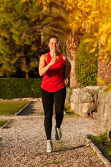 Fototapeta na wymiar Young Fit Woman Wearing Red Top And Leggings Doing Cardio Exercises In The Park With Stones Decoration And Trees On Summer Warm Day