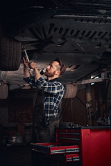 Bearded auto mechanic in a uniform repair the car's suspension with a wrench while standing under lifting car in repair garage. 