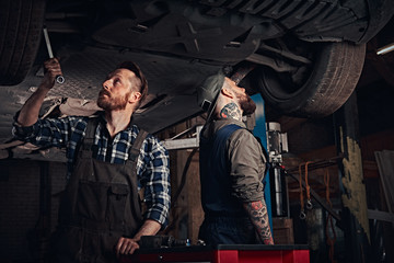 Fototapeta na wymiar Two bearded auto mechanic in a uniform repair the car's suspension while standing under lifting car in the repair garage. 
