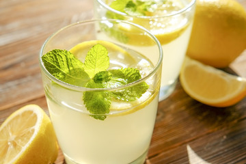 Two glasses full of ice cold refreshing lemonade beverage with mint leaves in sunlight on brown grunged wooden table, slices of ripe organic lemon, whole and halved. Background, copy space, close up.