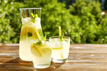 Lemonade pitcher and two glasses of cold refreshing citrus beverage with ice, lemon slices, mint leaves & straws on brown grunged wooden table, country side foliage background. Close up, copy space.