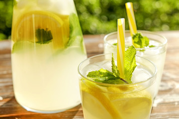 Lemonade pitcher and two glasses of cold refreshing citrus beverage with ice, lemon slices, mint leaves & straws on brown grunged wooden table, country side foliage background. Close up, copy space.