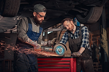 Fototapeta na wymiar Two bearded auto mechanic in a uniform and safety glasses working with an angle grinder while standing under lifting car in repair garage. 