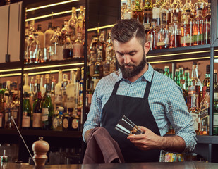 Stylish brutal barman is cleaning the glass with a cloth at bar counter background.