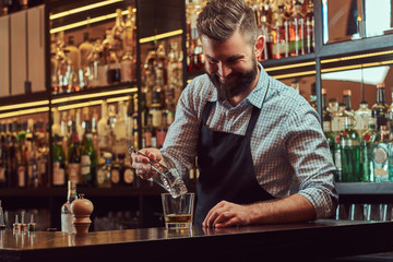 Stylish bearded barman in a shirt and apron splits ice to make a cocktail at bar counter background.