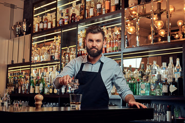 Stylish brutal barman in a shirt and apron makes a cocktail at bar counter background.