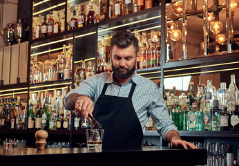 Stylish brutal barman in a shirt and apron makes a cocktail at bar counter background.