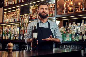 Stylish brutal bartender in a shirt and apron presents a bottle of exclusive alcohol at bar counter background.