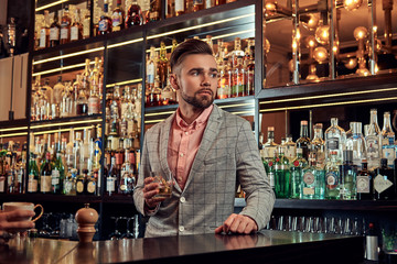 Stylish handsome male in an elegant suit holds a glass of whiskey at bar counter background.