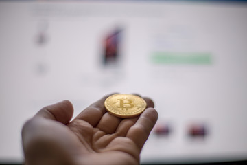 Hand holding Bitcoin coin offering to computer screen with store site on screen. Concept of virtual purchase with Bitcoin coin