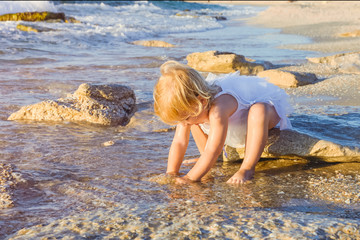 A cute little adorable toddler girl in white clothes playing with sand and shells on the empty beach on a warm sunny summer day. Holidays at sea. Family vacation. Selective focus, space for text.
