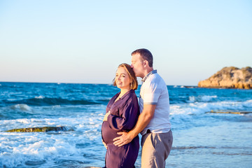 Portrait of happy love pregnant couple embracing each other and watching ahead to the sea during walk on the beach. Relax by the calm sea in sunshine. Family vacation, travelling. Selective focus.