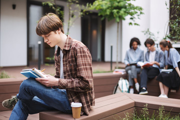 Young thoughtful man sitting on bench with coffee and writing in notebook while spending time in courtyard of university with students on background