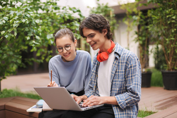 Couple of joyful students sitting on bench and working on laptop in courtyard of university. Young man and lady studying together outdoors