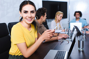 smiling young businesswoman with smartphone and her partners having discussion behind at modern office