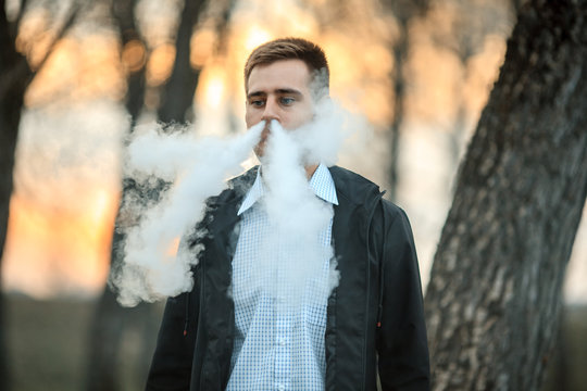 Vape teenager. Handsome young white guy in black jacket and checkered shirt vaping an electronic cigarette among the trees in the park at sunset. Lifestyle.
