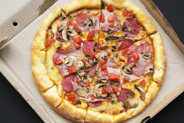 Top view, pizza with bacon, tomatoes, salami, cheese and mushrooms in a cardboard box on dark background. From above, flat lay.