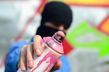 A young graffiti artist in a blue jacket and black mask is holding a can of paint in front of him...