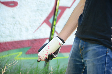 A young hooligan with a spray can stands against a concrete wall with graffiti paintings. Illegal...