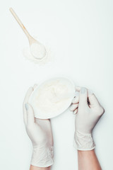 cropped shot of woman in latex gloves with plate with clay mask on surface with spoon and clay powder