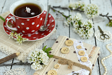 Fototapeta na wymiar Red cup of tea with hand made diary and plum tree branches in blossom. Spring still life, beautiful rural background with seasonal flowers in vintage style