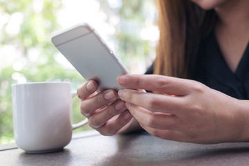 Closeup image of a woman holding and using smartphone with coffee cup in cafe
