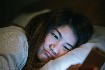 Young asian woman on bed late at night texting using mobile phone sleepy and tired in internet communication overuse and smartphone addiction with boring emotion.