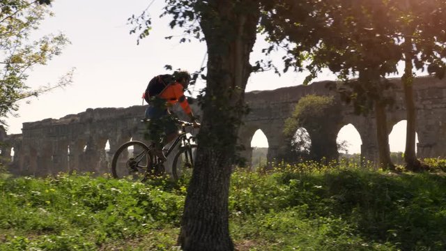 Cyclist riding bike in front of ancient roman aqueduct in a park. Young attractive athletic man with orange sportswear and backpack in parco degli acquedotti in Rome