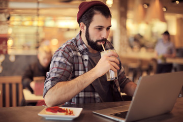 Handsome trendy hipster with glass of coffee drink using laptop in cafe