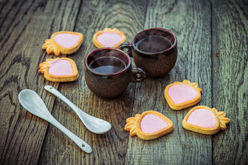 Obraz na płótnie Canvas Ceramic cups with hot freshly brewed coffee, strawberry shape cookies with pink cream and white spoons on a wooden table. Sweet snack or breakfast.