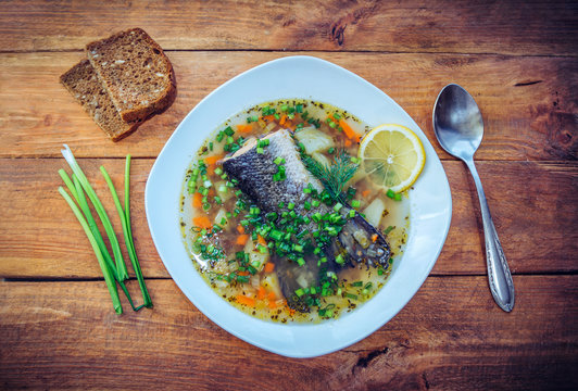 Fish soup with salmon tail, lemon slice and cut green onion in a plate, bread with sunflower seeds and spoon on the wooden table