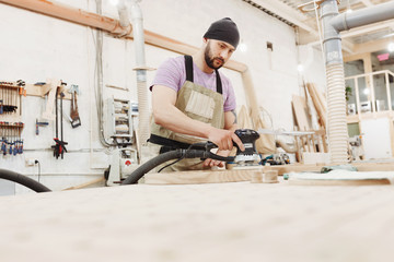 A young bearded male joiner uses random orbit sander, processes wooden products to produce furniture in carpenter workshop