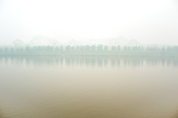 The shore of the pond, trees and village houses in a strong fog. Summer day.
