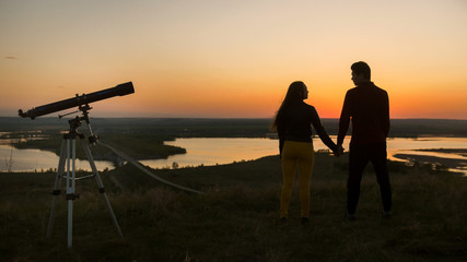 Fototapeta na wymiar Telescope stands in front of couple holding hands on the hill at the amazing sunset