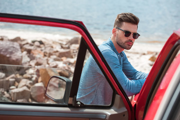 stylish male traveler standing at car near the sea during road trip