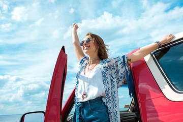 excited young woman in sunglasses standing near car on road trip