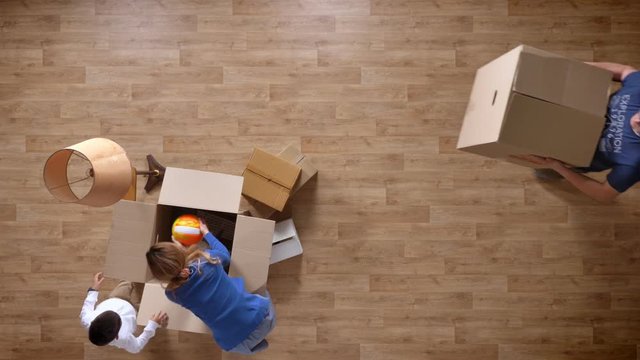 Young mother and son unpacking box, father coming with box, boy running with ball top shot parquet wooden floor