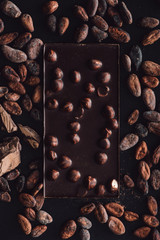 top view of chocolate bar with hazelnuts surrounded by cocoa beans on dark surface