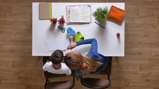 Young mother painting with son, sitting behind table, kid doing homework top shot parquet wooden floor