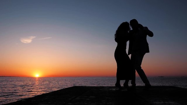 Silhouettes of young spanish couple against sunset sea background. Man and woman dancing latin american sensual dance: bachata, salsa or kizomba. Slow motion.