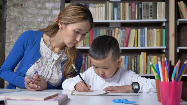 Asain young mother helping son with study, women speaking to kid, sitting behind table, asian kid doing homework, book shelves background
