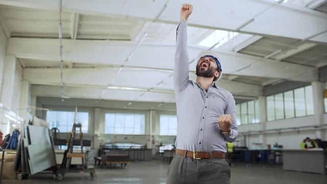 Portrait of adult bearded man, head director or engineer, wearing suit and helmet rejoicing with clenching fists after making business deal in big factory slow motion