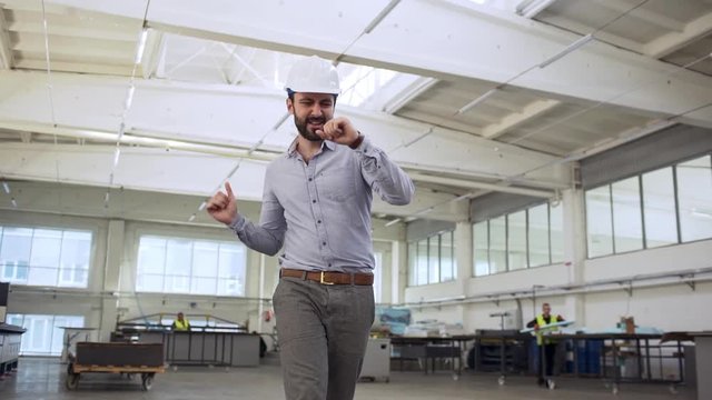 Portrait of adult happy man, head director or engineer, wearing suit and helmet rejoicing and dancing while walking though manufacture slow motion