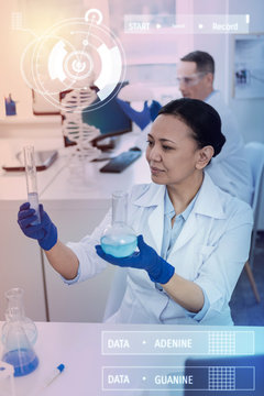 Holding glass bottles. Calm professional chemist sitting in a modern lab and carefully holding two glass bottles with blue liquid in them